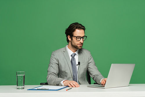 bearded news anchor in eyeglasses and suit using laptop isolated on green