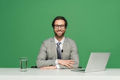 bearded news anchor in eyeglasses and suit smiling while sitting near laptop isolated on green