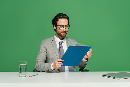 smiling news anchor in eyeglasses and suit holding clipboard near laptop isolated on green