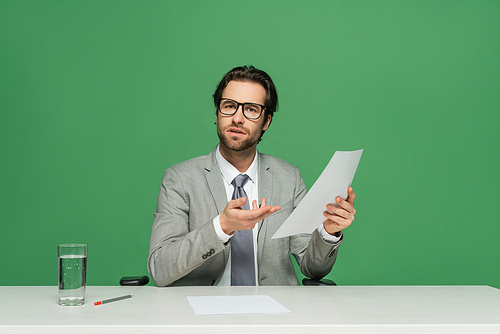 confused news anchor in eyeglasses and suit pointing with hand at blank paper isolated on green