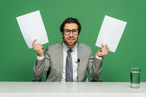 emotional news anchor in eyeglasses and suit holding blank papers isolated on green