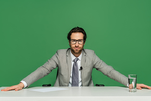 broadcaster in eyeglasses and grey suit sitting at desk isolated on green