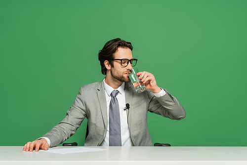 broadcaster in eyeglasses sitting at desk and drinking  isolated on green