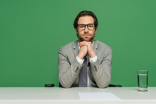 broadcaster in eyeglasses and grey suit sitting with clenched hands at desk isolated on green