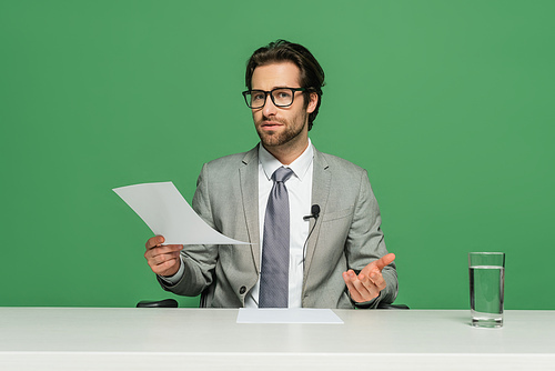 news broadcaster in eyeglasses and suit sitting at desk and holding paper isolated on green