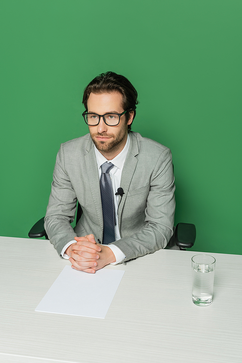 overhead view of news broadcaster in eyeglasses and suit sitting with clenched hands at desk isolated on green