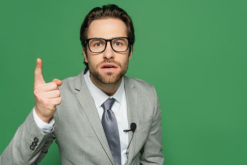 emotional newscaster in eyeglasses and suit pointing with finger isolated on green