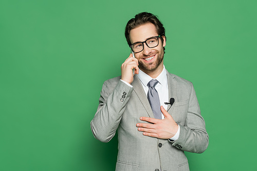 cheerful reporter in eyeglasses and suit talking on smartphone on green background