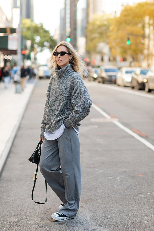 full length of stylish woman in sunglasses and grey outfit holding handbag while posing on street of New York city