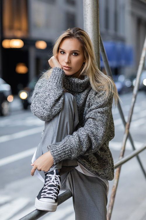 young blonde woman in trendy winter outfit and footwear sitting on urban street in New York
