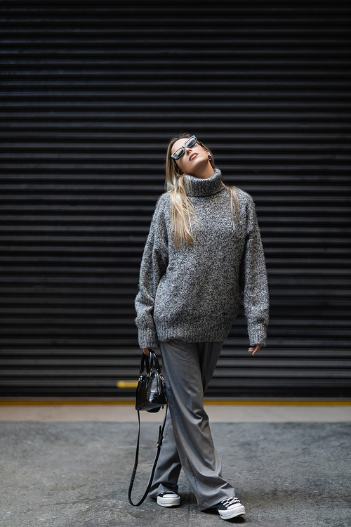 full length of blonde woman in winter sweater and sunglasses walking with handbag on urban street