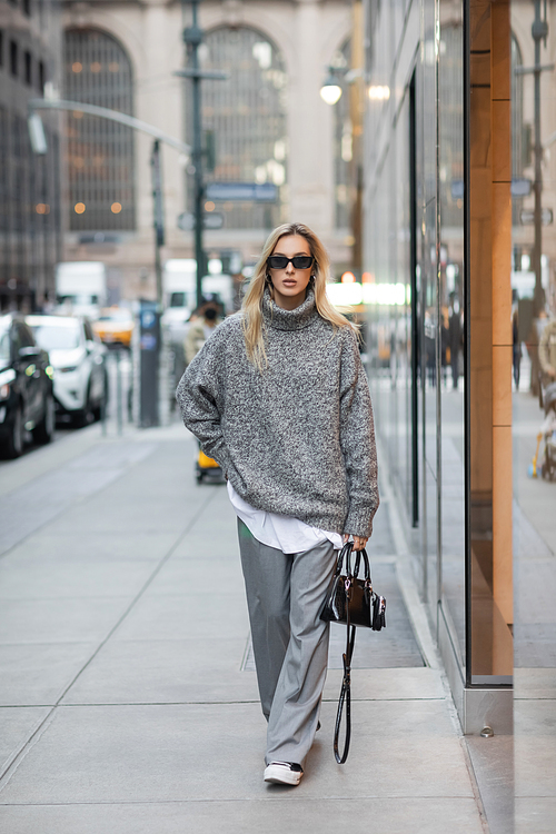 full length of blonde woman in winter sweater and sunglasses walking with handbag on urban street in New York