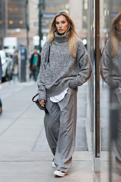 full length of blonde woman in winter sweater holding sunglasses and handbag on urban street in New York
