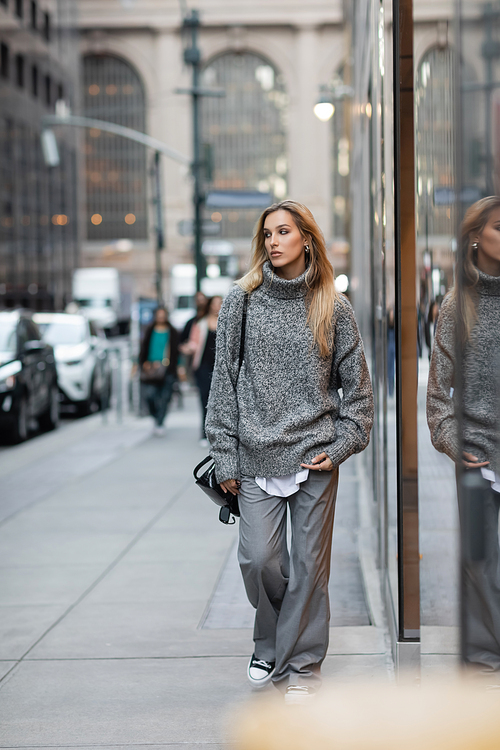 full length of blonde woman in winter sweater holding sunglasses and handbag while walking on urban street in New York