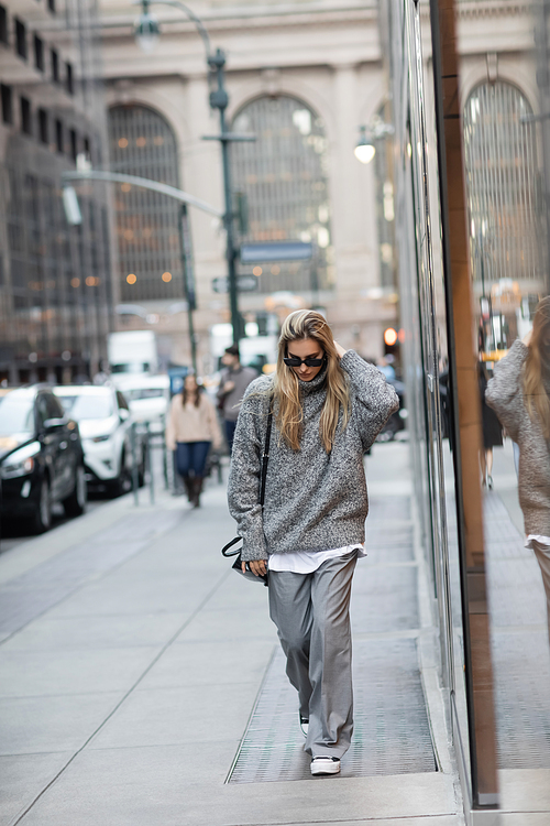 full length of woman in winter sweater and sunglasses walking with handbag on urban street in New York
