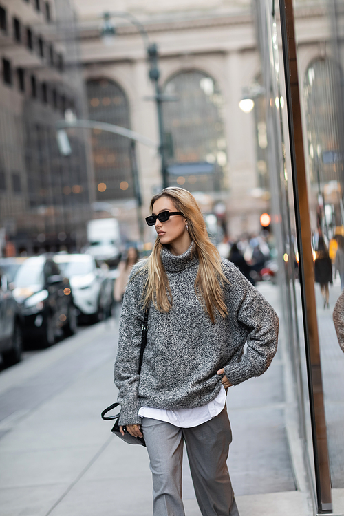 pretty woman in winter sweater and sunglasses standing with hand on hip on urban street in New York