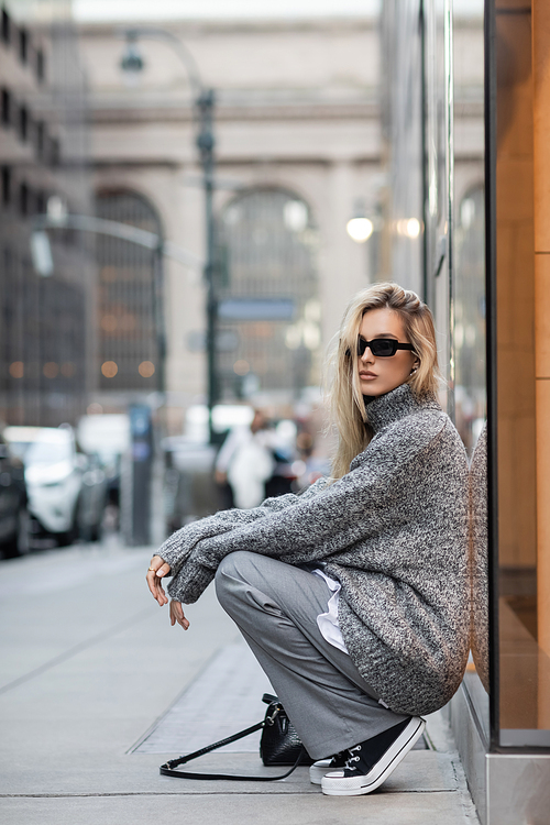 full length of young woman in winter sweater and sunglasses sitting near handbag on urban street in New York city
