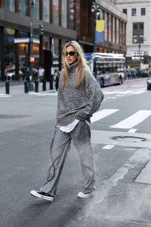 full length of blonde woman in stylish knitted sweater and sunglasses walking on urban street in New York