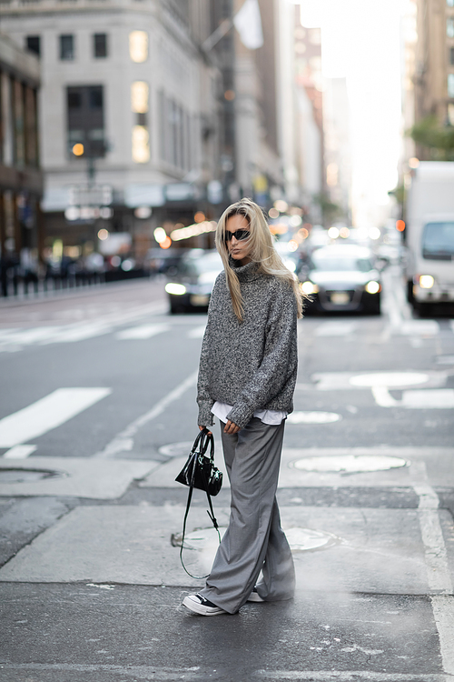 full length of blonde woman in knitted sweater and sunglasses walking with trendy handbag on urban street in New York