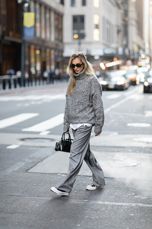 full length of blonde woman in knitted sweater and sunglasses walking with trendy handbag near cars in New York