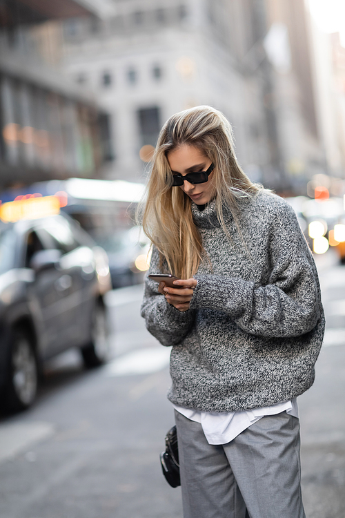 concentrated woman in winter sweater and sunglasses using smartphone on urban street in New York city