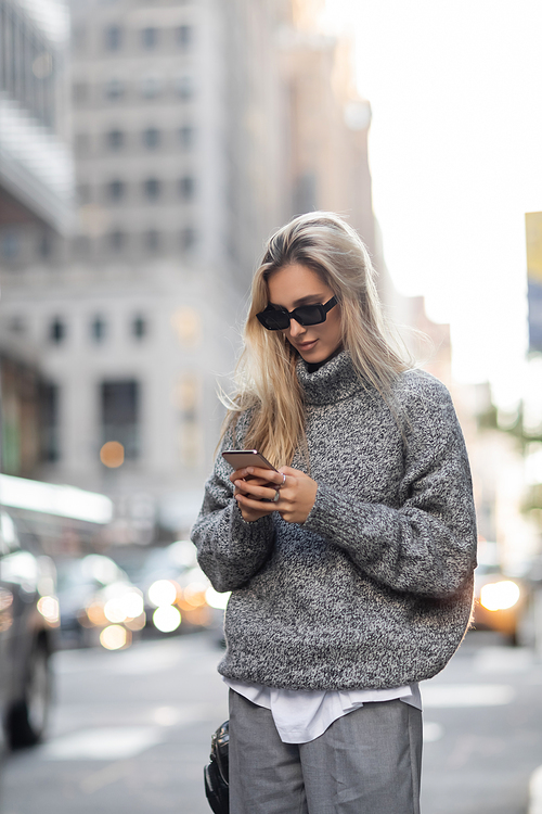 blonde woman in knitted sweater and sunglasses using smartphone on urban street in New York city