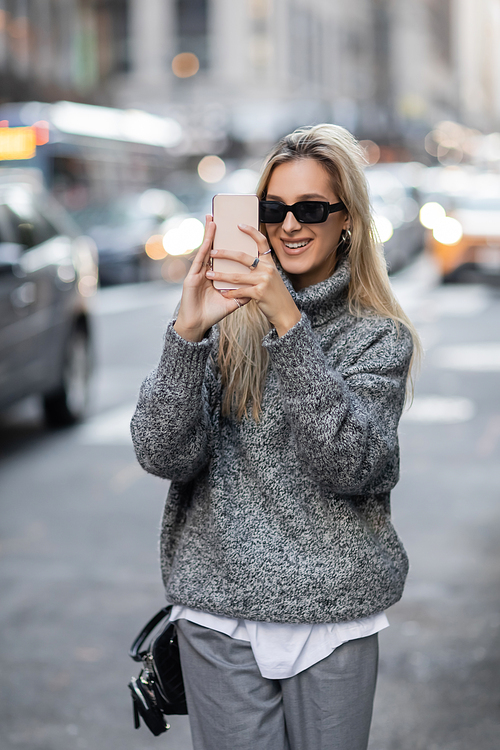 happy young woman in winter sweater and sunglasses taking photo on smartphone in New York city