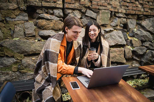 Positive multiethnic couple in blanket using laptop on terrace of cafe