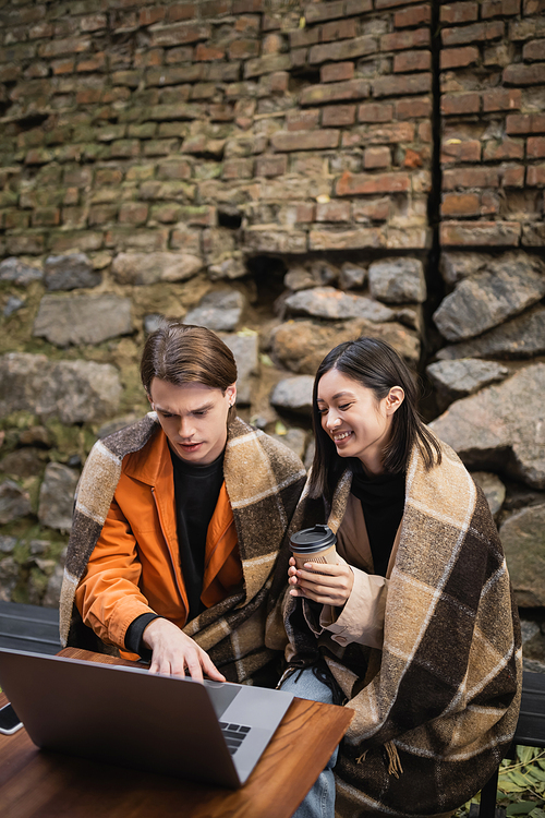Cheerful asian woman holding coffee to go near boyfriend using laptop on terrace of cafe