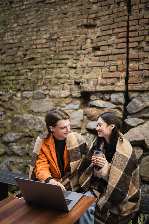 Multiethnic couple with blanket and coffee talking near devices in outdoor cafe