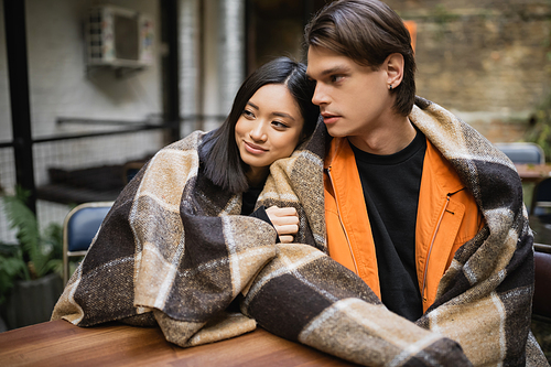 Smiling asian woman in blanket hugging boyfriend on terrace of cafe