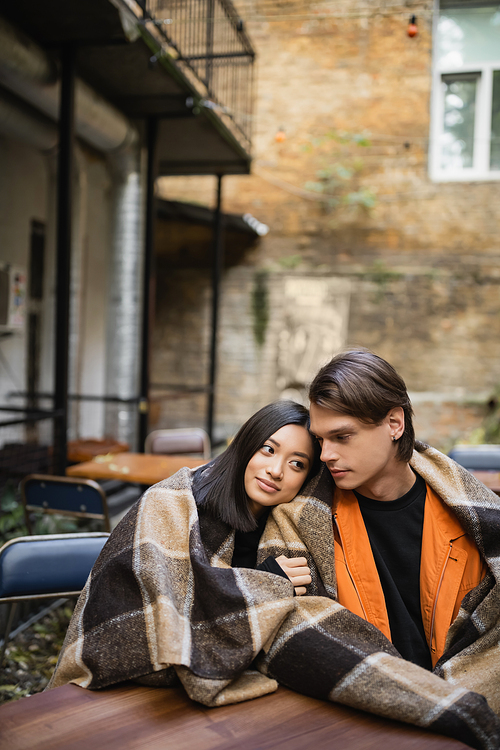 Young asian woman in blanket hugging boyfriend on terrace of cafe