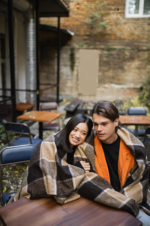 Cheerful asian woman in blanket hugging boyfriend on terrace of cafe