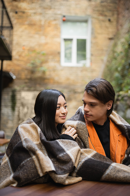 Young multiethnic couple in blanket looking at each other in outdoor cafe