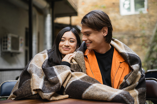 Smiling man looking at asian girlfriend in blanket in outdoor cafe