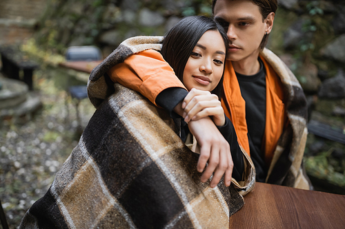 Young man hugging asian girlfriend in blanket on terrace of cafe