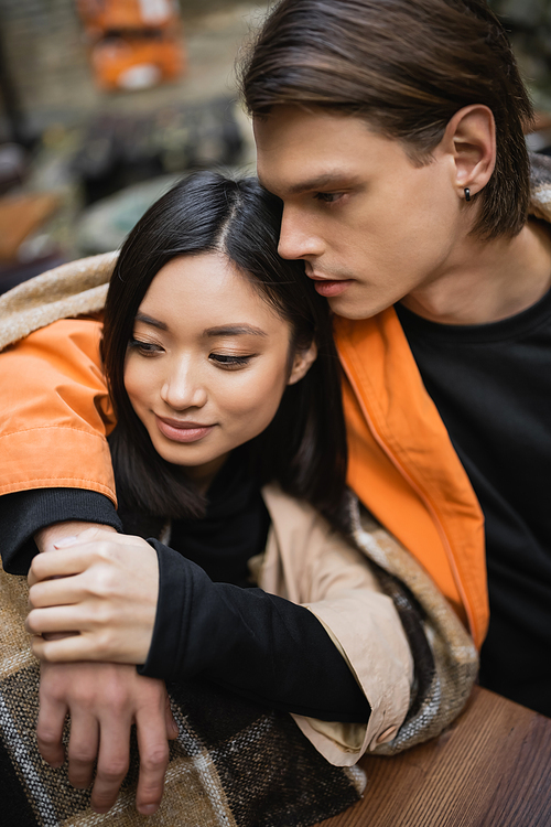 Brunette man hugging young asian girlfriend in blanket on cafe terrace