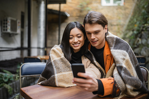 Smiling interracial couple in blanket using smartphone in outdoor cafe