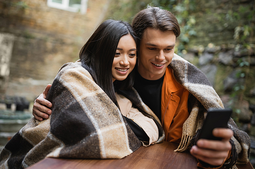 Young man hugging asian girlfriend in blanket while using blurred smartphone on cafe terrace