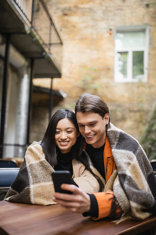 Positive interracial couple in blanket using cellphone on terrace of cafe