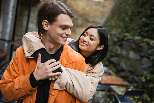 Smiling asian woman in trench coat embracing boyfriend on terrace of cafe