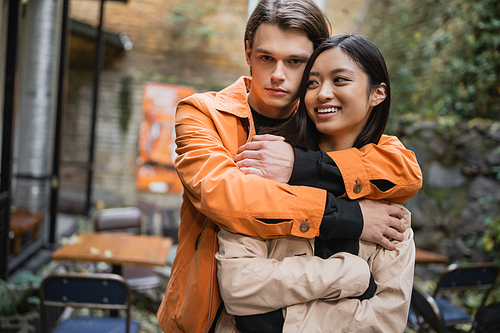 Young man hugging cheerful asian girlfriend on terrace of cafe