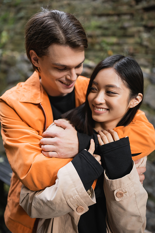 Smiling man hugging cheerful and stylish girlfriend on terrace of cafe