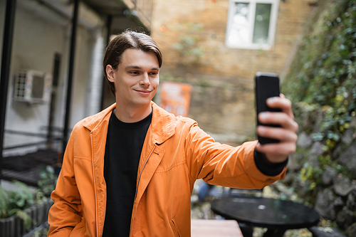 Smiling man in jacket taking selfie on smartphone on terrace of cafe