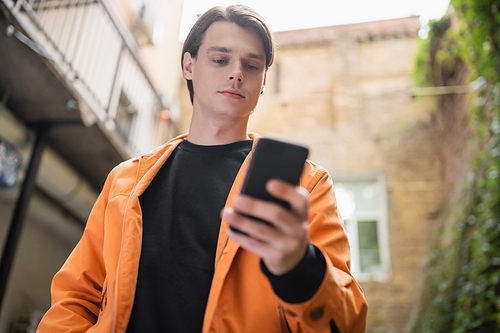 Low angle view of young man in jacket using blurred smartphone on cafe terrace