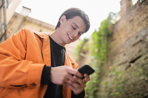 Low angle view of smiling man in jacket using mobile phone outdoors
