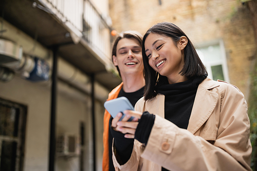 Smiling asian woman in trench coat using smartphone near blurred boyfriend on terrace of cafe