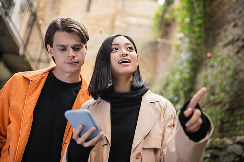 Low angle view of young asian woman using smartphone and pointing with finger near boyfriend outdoors