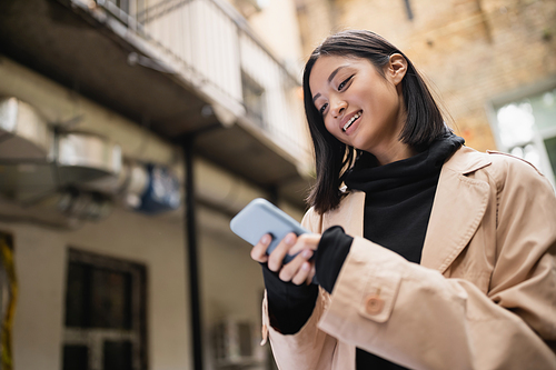 Low angle view of smiling asian woman in trench coat using smartphone outdoors