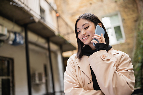 Cheerful asian woman in trench coat talking on smartphone in outdoor cafe